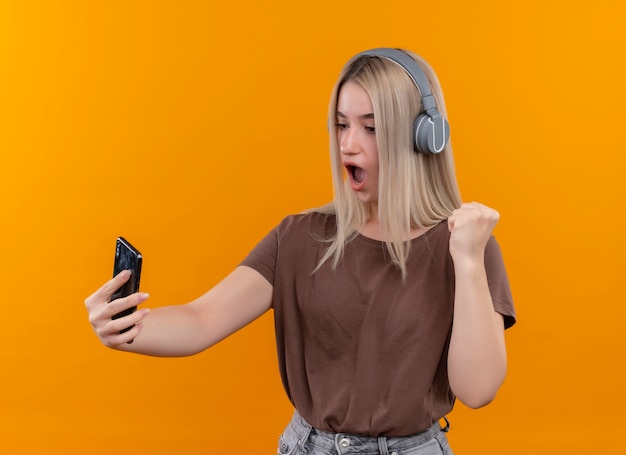 Verrast jong blondemeisje die hoofdtelefoons dragen die mobiele telefoon houden die het met opgeheven vuist op geïsoleerde oranje muur bekijken