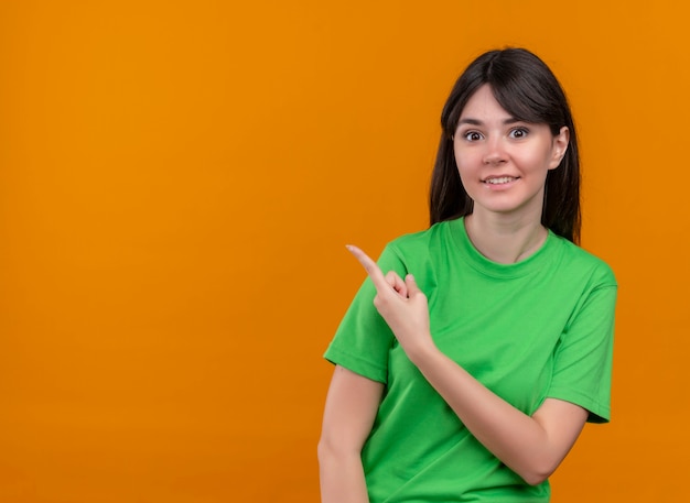 Verrast jong kaukasisch meisje in groen shirt wijst naar de zijkant en kijkt naar de camera op geïsoleerde oranje achtergrond met kopie ruimte