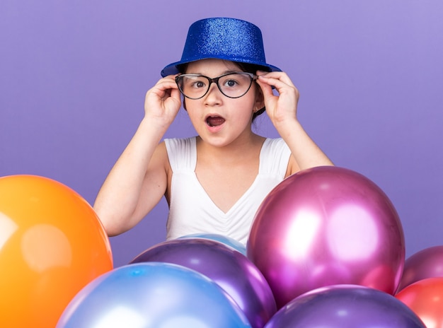 verrast jong Kaukasisch meisje met blauwe feestmuts in optische bril permanent met helium ballonnen geïsoleerd op paarse muur met kopie ruimte