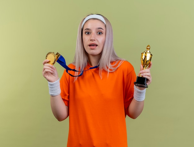 Verrast jong kaukasisch sportief meisje met beugels met hoofdband en polsbandjes houdt gouden medaille en winnaarbeker