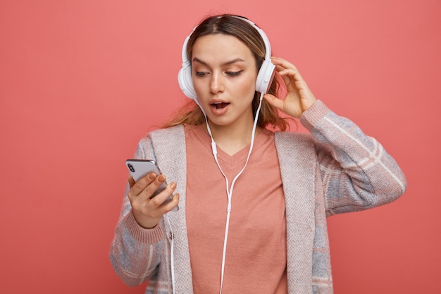 Verrast jong meisje dragen en koptelefoon houden en kijken naar mobiele telefoon