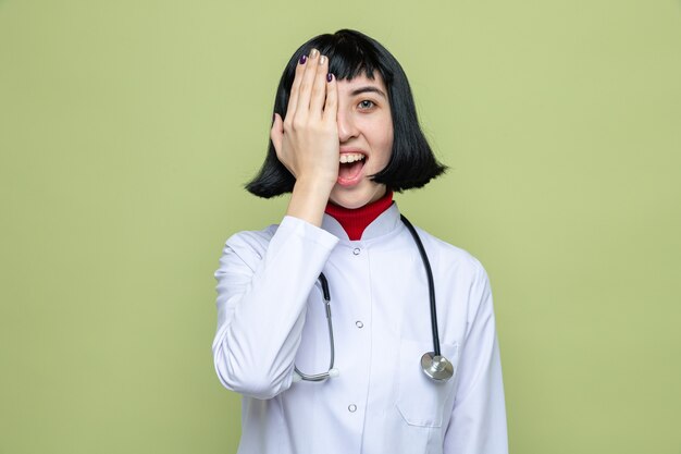 Verrast jong, mooi kaukasisch meisje in doktersuniform met een stethoscoop die haar oog bedekt met de hand en naar de voorkant kijkt