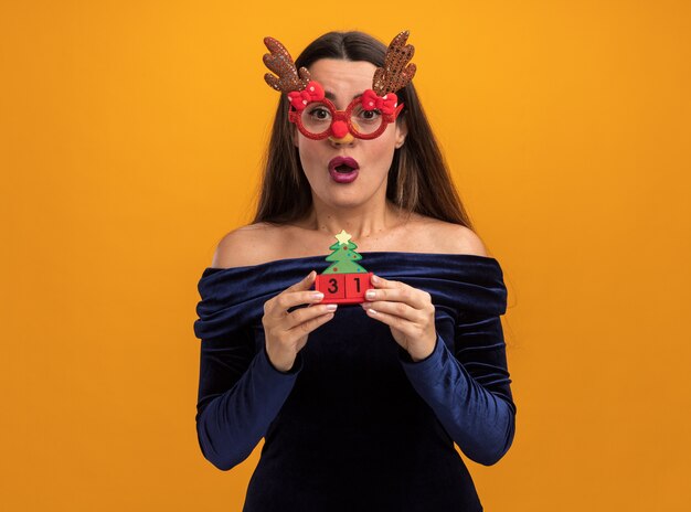 Verrast jong mooi meisje die blauwe kleding en Kerstmisglazen dragen die stuk speelgoed houden dat op oranje achtergrond wordt geïsoleerd