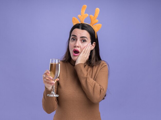 Verrast jong mooi meisje dragen bruine trui met kerst haar hoepel glas champagne hand zetten wang geïsoleerd op blauwe achtergrond