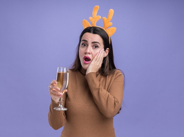 Verrast jong mooi meisje dragen bruine trui met kerst haar hoepel glas champagne hand zetten Wang geïsoleerd op blauwe achtergrond