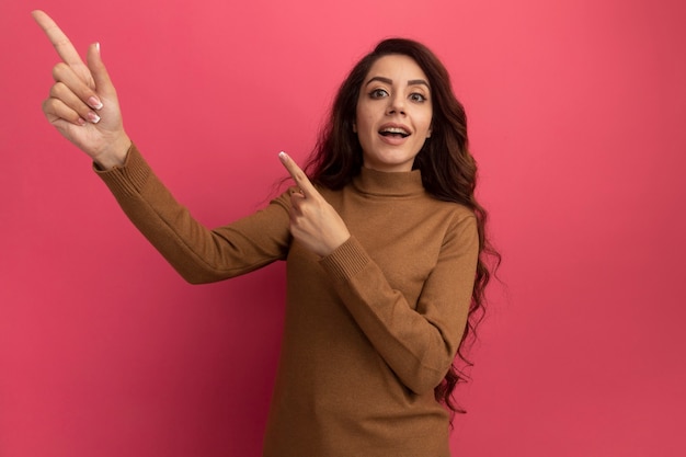 Verrast jong mooi meisje met coltrui wijst naar kant geïsoleerd op roze muur