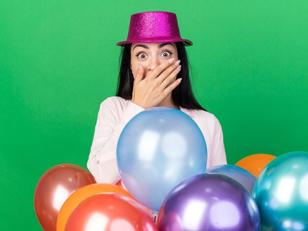 Verrast jong mooi meisje met een feestmuts die achter ballonnen staat en de mond bedekt met de hand?