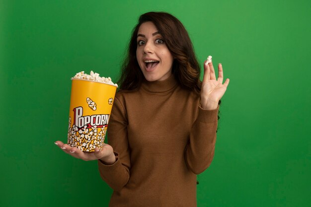 Verrast jong mooi meisje met emmer popcorn met popcorn vrede geïsoleerd op groene muur