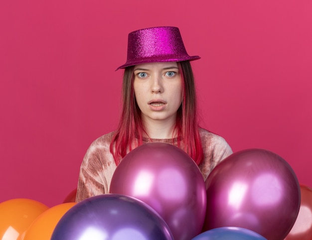 Verrast jong mooi meisje met feestmuts achter ballonnen geïsoleerd op roze muur
