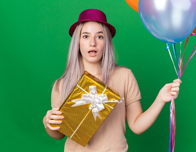 Verrast jong mooi meisje met feestmuts en beugels met ballonnen met geschenkdoos