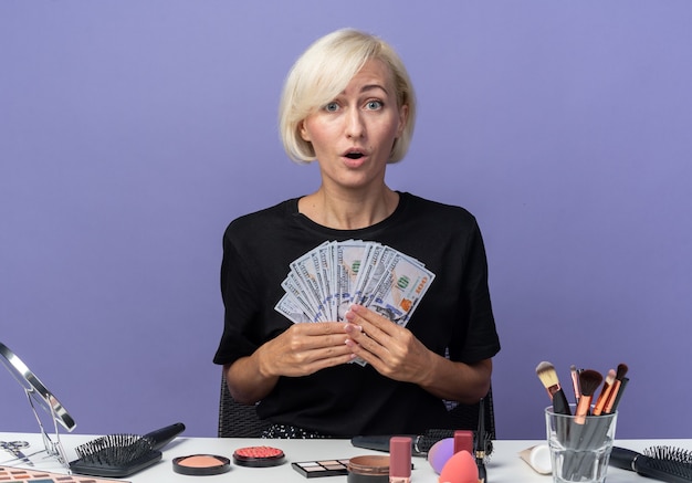 Verrast jong mooi meisje zit aan tafel met make-up tools met contant geld geïsoleerd op blauwe achtergrond