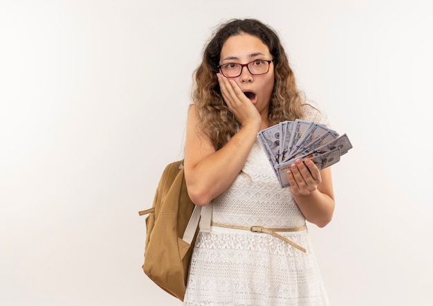 Verrast jong mooi schoolmeisje die glazen en achterzak dragen die geld met hand op gezicht houden dat op witte muur wordt geïsoleerd