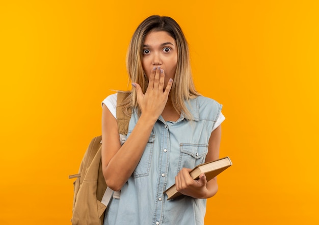 Verrast jong mooi studentenmeisje die het boek van de achterzakholding dragen en hand op mond zetten die op oranje muur wordt geïsoleerd