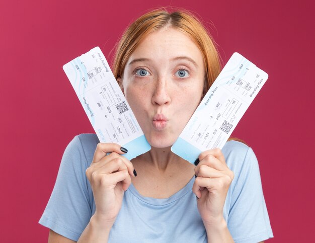 Verrast jong roodharig gembermeisje met sproeten die vliegtickets dicht bij het gezicht op roze houden