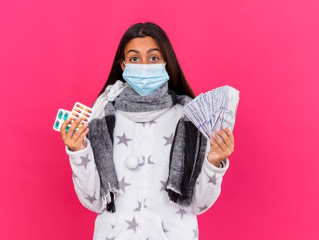Verrast jong ziek meisje dat medisch masker met de pillen van de sjaalholding met contant geld draagt die op roze wordt geïsoleerd