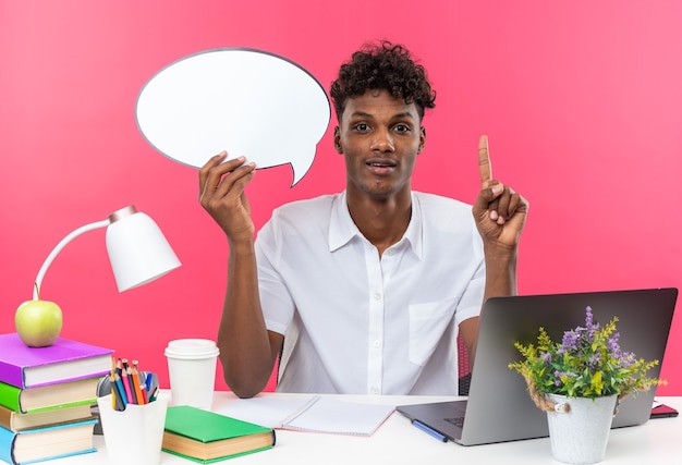 Verrast jonge Afro-Amerikaanse student zit aan een bureau met schoolhulpmiddelen die een tekstballon vasthouden en naar boven wijzen