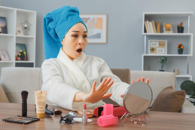 Verrast jonge aziatische vrouw met handdoek op haar hoofd zittend aan de kaptafel in het interieur kijken naar haar nagels verward zijn ochtend make-up routine