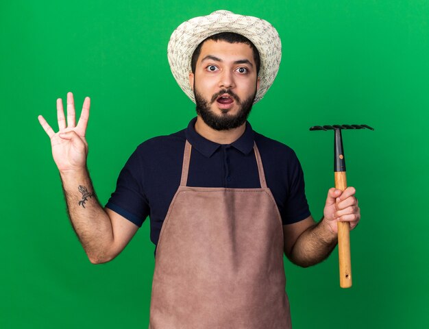 verrast jonge blanke mannelijke tuinman dragen tuinieren hoed bedrijf hark en gebaren vier met vingers geïsoleerd op groene muur met kopie ruimte