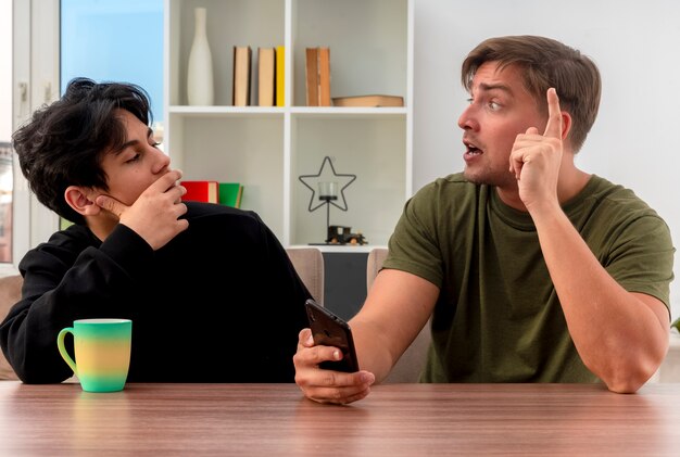 Verrast jonge blonde knappe man wijst naar telefoon zittend aan tafel en kijken naar elkaar met zelfverzekerde jonge brunette knappe jongen hand op kin zetten in woonkamer