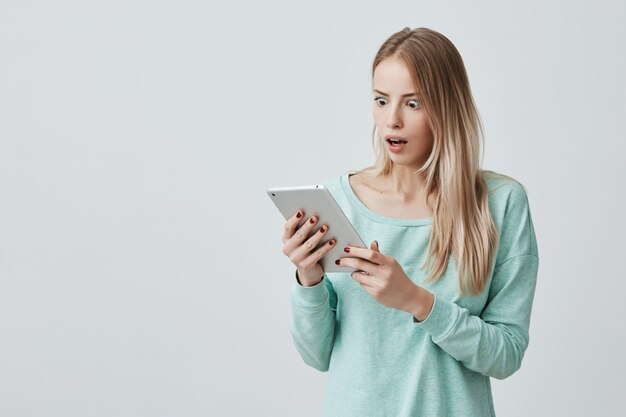 Verrast jonge blonde vrouw in top met lange mouwen met wijd open mond in shock en verbazing kijken naar scherm van tablet