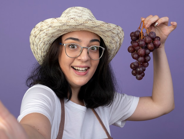 Gratis foto verrast jonge brunette vrouwelijke tuinman in optische bril en uniform dragen tuinieren hoed houdt druiven kijken voorzijde geïsoleerd op paarse muur