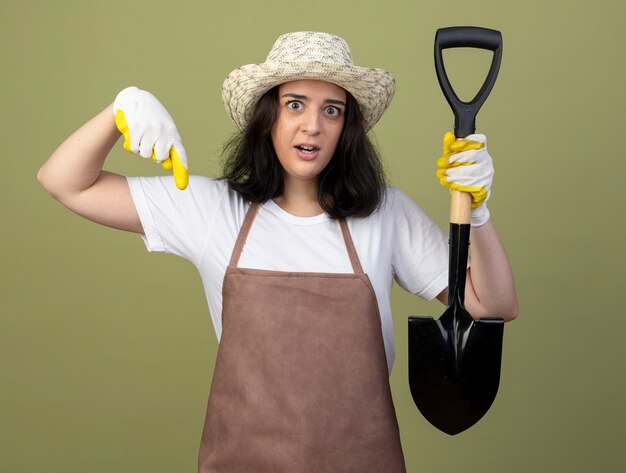 Verrast jonge brunette vrouwelijke tuinman in uniform dragen tuinieren hoed en handschoenen houdt schop en wijst naar beneden geïsoleerd op olijfgroene muur