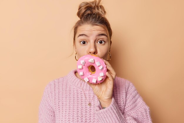 Verrast jonge Europese vrouw bedekt mond met heerlijke grote smakelijke donut heeft suikerverslaving haar gekamd in broodje draagt gebreide trui geïsoleerd over beige achtergrond. Zoete tand concept