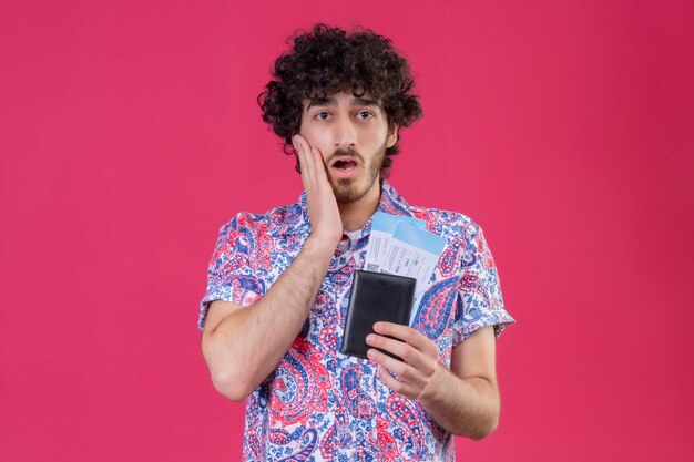Verrast jonge knappe gekrulde reiziger man met portemonnee en vliegtuigtickets met hand op wang op geïsoleerde roze muur met kopie ruimte