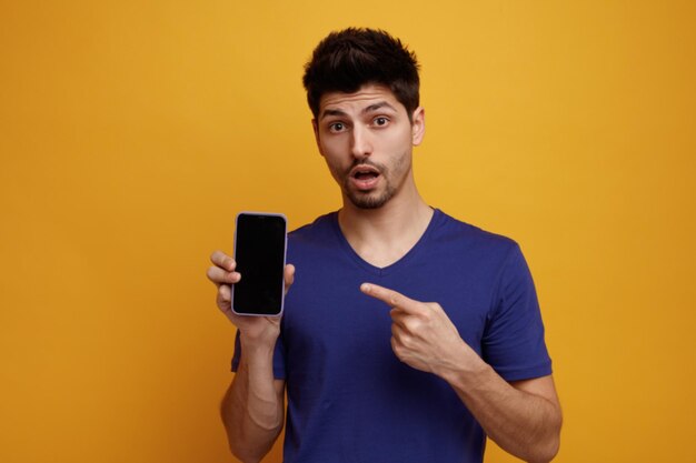 Verrast jonge knappe man die naar de camera kijkt met een mobiele telefoon die ernaar wijst op een gele achtergrond