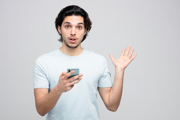 verrast jonge knappe man met mobiele telefoon kijken naar camera met lege hand geïsoleerd op een witte achtergrond