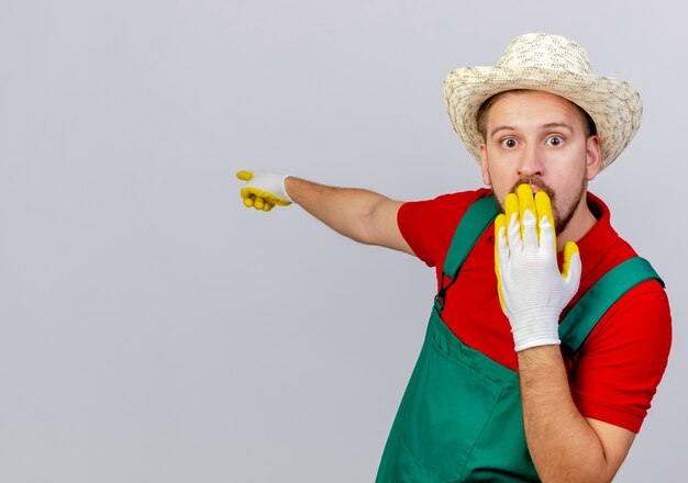 Verrast jonge knappe Slavische tuinman in uniform met tuinhandschoenen en hoed op zoek houden hand op mond wijzend achter geïsoleerd