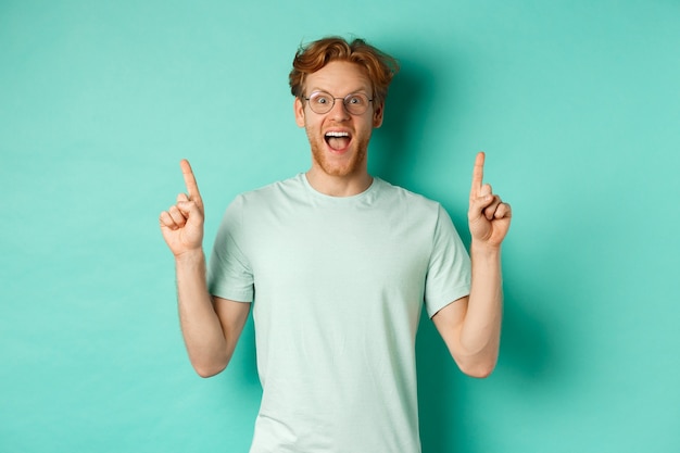 Verrast jonge man met rood haar, bril en t-shirt dragen, hijgend van ontzag en wijzende vingers naar promo deal, staande over mint achtergrond