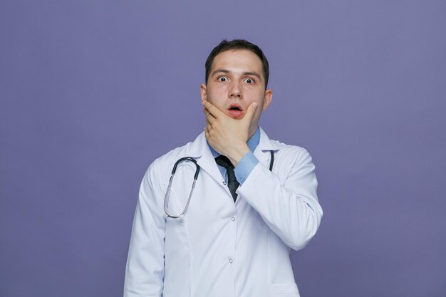 Verrast jonge mannelijke arts dragen medische mantel en stethoscoop rond de nek camera kijken terwijl hand op kin geïsoleerd op paarse achtergrond