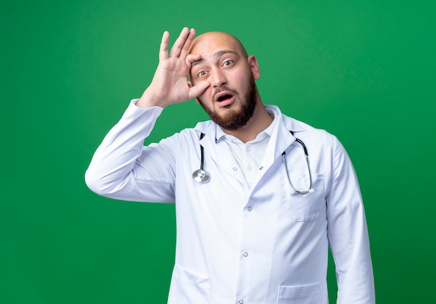 Verrast jonge mannelijke arts medische gewaad en stethoscoop dragen oog openen met hand geïsoleerd op groen