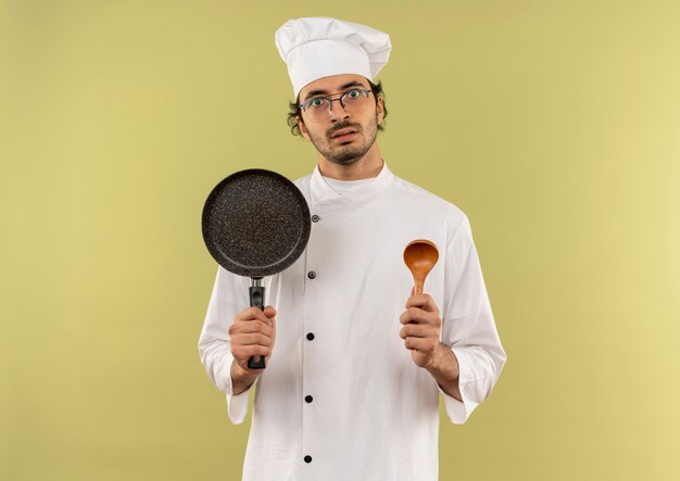 verrast jonge mannelijke kok die uniforme chef-kok en glazen draagt die koekenpan en lepel houdt