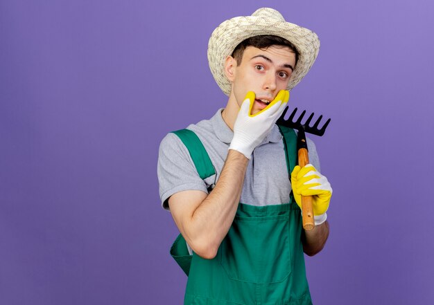 Verrast jonge mannelijke tuinman met tuinhoed en handschoenen houdt hark vast