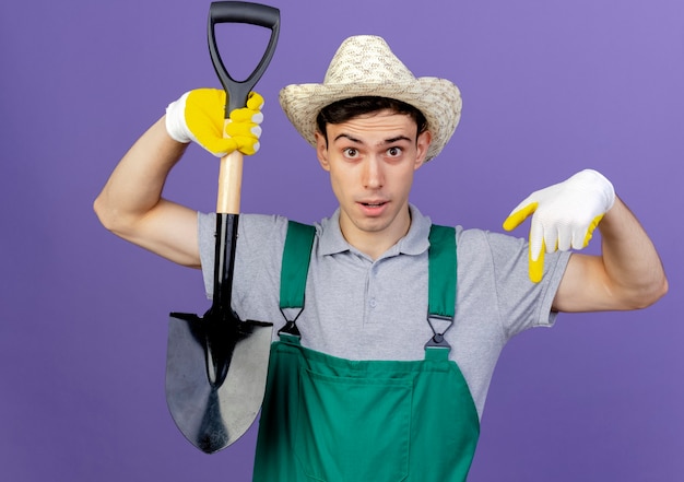 Verrast jonge mannelijke tuinman met tuinhoed en handschoenen houdt schop vast