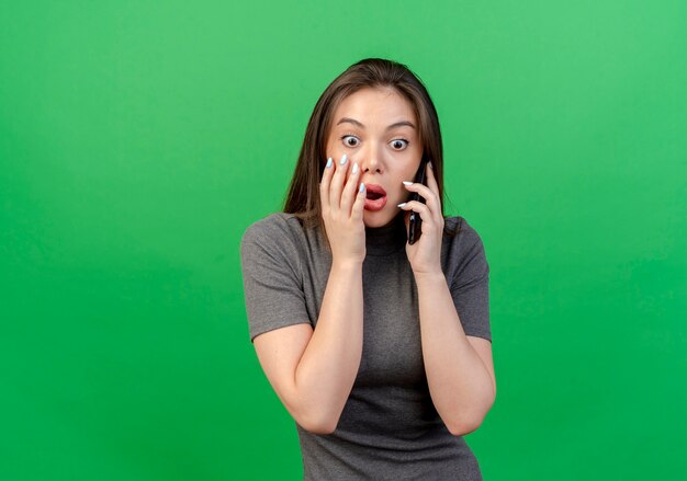 Verrast jonge mooie vrouw op zoek rechtstreeks praten aan de telefoon aanraken gezicht geïsoleerd op groene achtergrond met kopie ruimte