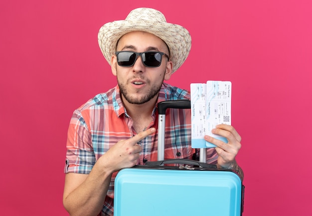 Gratis foto verrast jonge reiziger man met stro strand hoed in zonnebril houden en wijzend op vliegtickets staan achter koffer geïsoleerd op roze muur met kopie ruimte