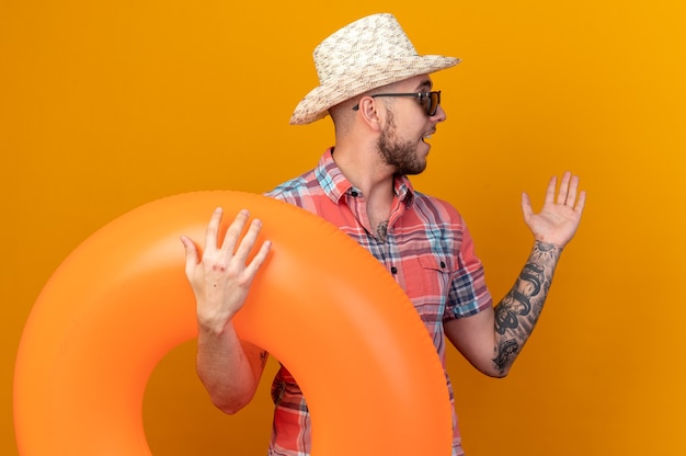 Verrast jonge reiziger man met stro strand hoed in zonnebril met zwemring kijkend naar kant geïsoleerd op oranje muur met kopie ruimte