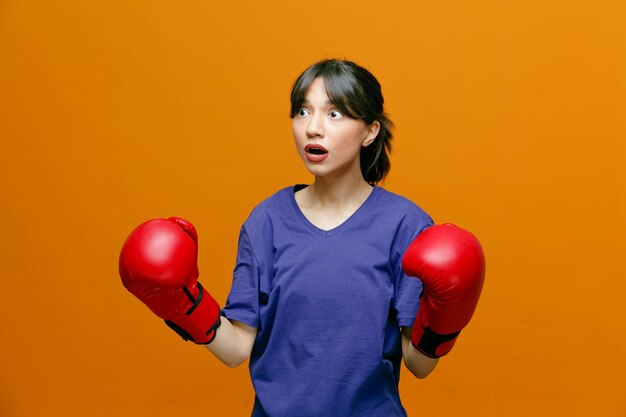Verrast jonge sportieve vrouw met t-shirt en bokshandschoenen die naar de zijkant kijkt en vuisten in de lucht houdt geïsoleerd op een oranje achtergrond
