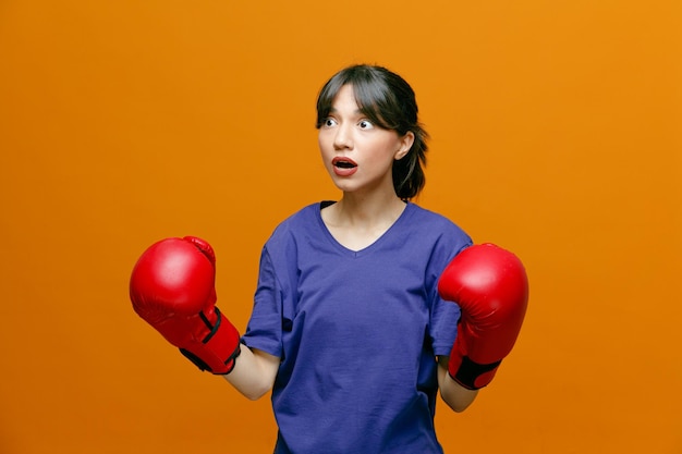 Verrast jonge sportieve vrouw met t-shirt en bokshandschoenen die naar de zijkant kijkt en vuisten in de lucht houdt geïsoleerd op een oranje achtergrond