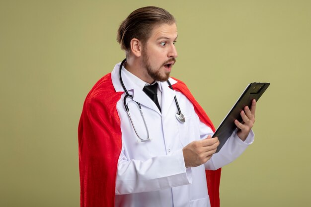 Verrast jonge superheld man dragen stethoscoop met medische gewaad houden en kijken naar klembord geïsoleerd op olijfgroene achtergrond