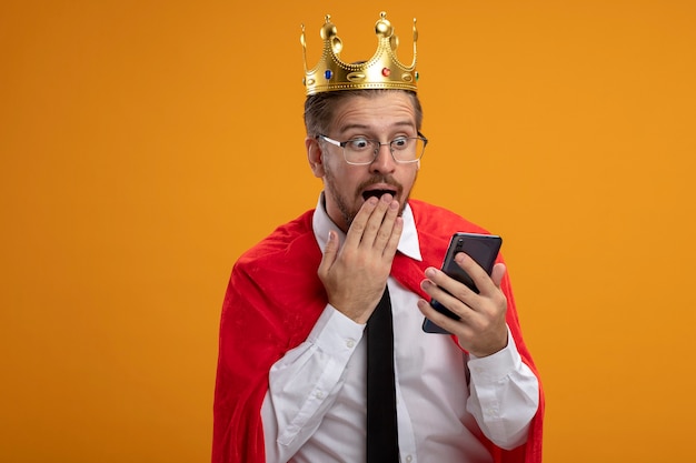 Verrast jonge superheld man stropdas en kroon met bril houden en kijken naar telefoon bedekt mond met hand geïsoleerd op een oranje achtergrond