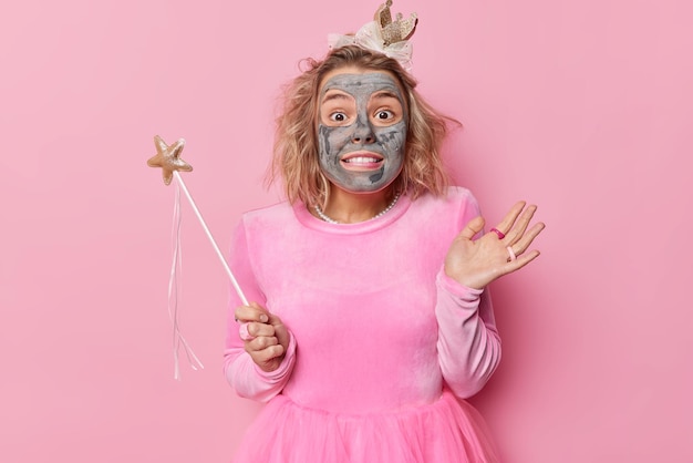 Verrast jonge vrouw entertaint gasten op feestje past schoonheid klei masker draagt jurk houdt toverstaf klaar om uw wensen en dromen te vervullen geïsoleerd over roze achtergrond Magische fee binnen