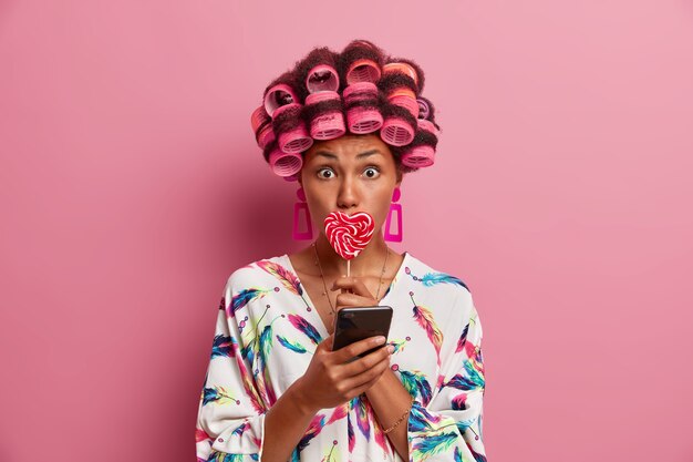 Verrast jonge vrouw gebruikt haarrollers om mooi kapsel te creëren, bedekt mond met lolly, gebruikt smartphone voor online communicatie, draagt zijden badjas, poseert tegen roze muur