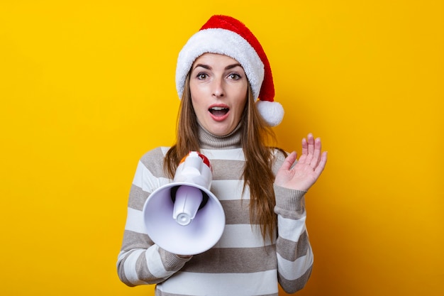 Verrast jonge vrouw in kerstman hoed met een megafoon op een gele achtergrond.