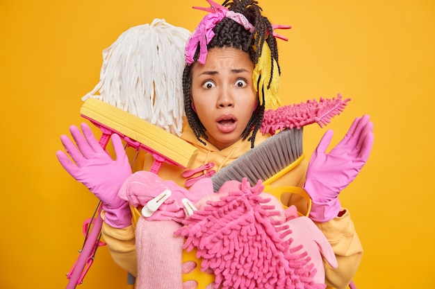 Gratis foto verrast jonge vrouw met donkere huid en dreadlocks poseert in de buurt van een emmer vol schoonmaakspullen