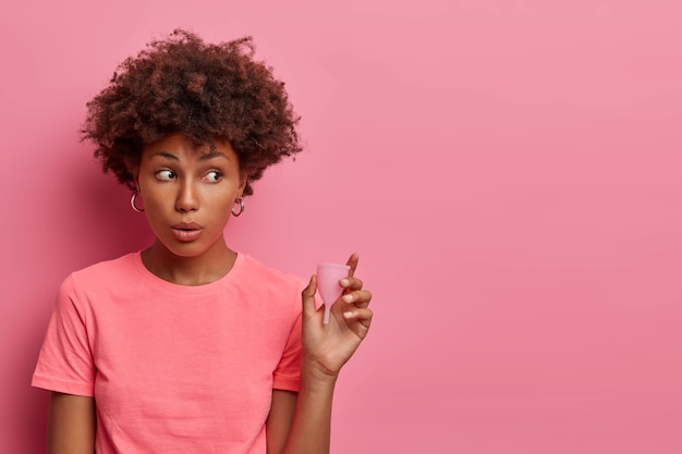 Verrast jonge vrouw vormt met apparaat voor vrouwelijke hygiëne dat tijdens de menstruatie in de vagina wordt ingebracht. vrouw houdt menstruatiecup vast om menstruatievloeistof op te vangen en te voorkomen dat het op kleding lekt