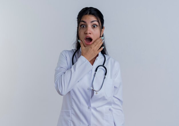 Verrast jonge vrouwelijke arts die medische gewaad en stethoscoop draagt die hand op geïsoleerde kin zetten